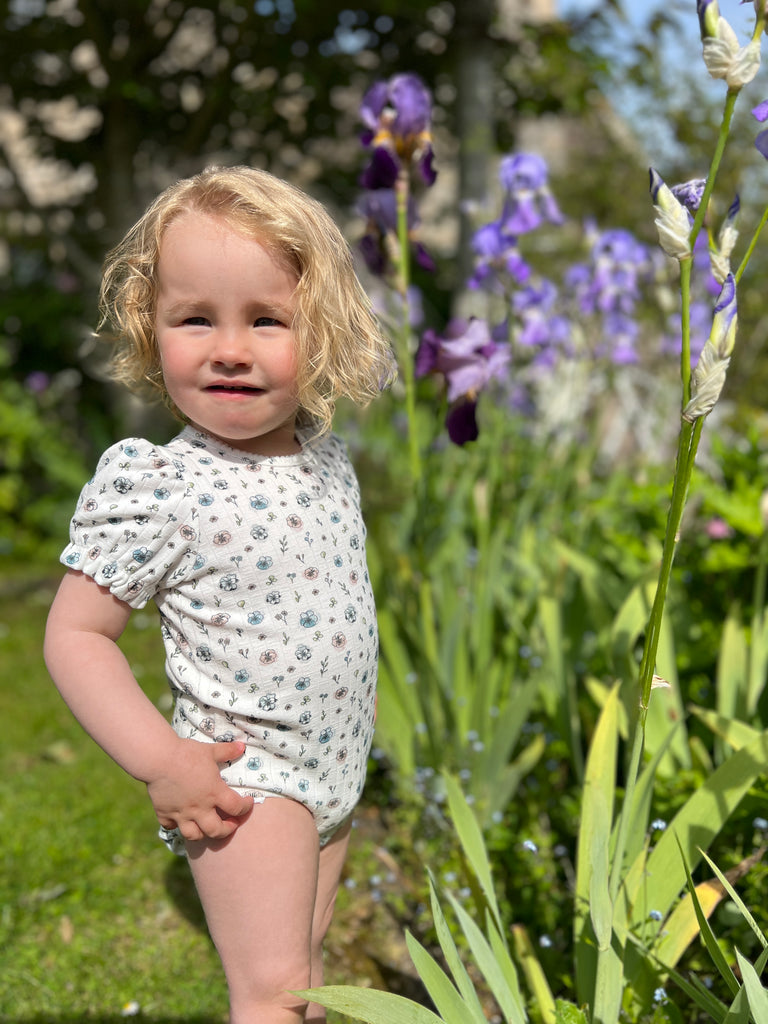 toddler standing in sunny garden wearing short sleeved white ribbed jersey onesie with  flower print
