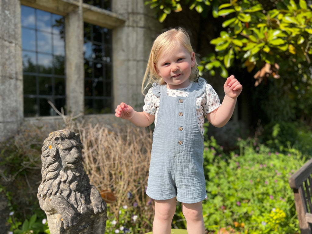 girl standing in front of old stone house on sunny day wearing blue shortie overalls with frill shoulder straps and buttons down the front with flower print tee underneath