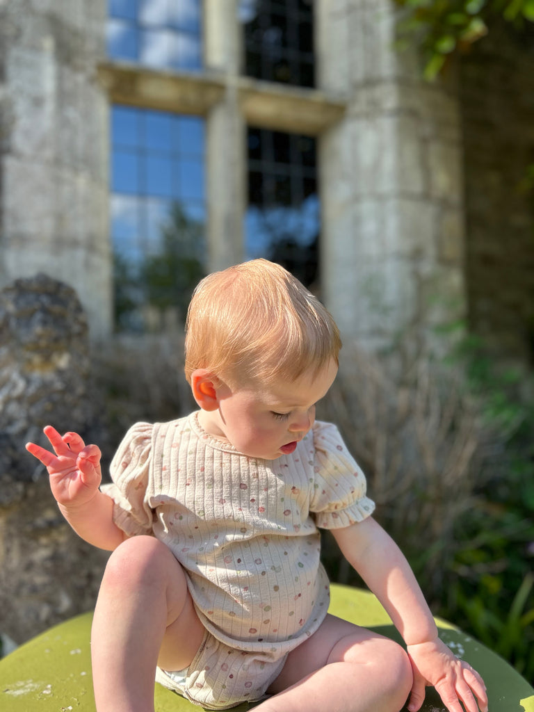baby sat in sunny garden in front of old stone house wearing short sleeved cream ribbed jersey onesie with pastel dot print