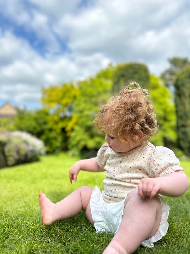 toddler in sunny garden wearing white gauze shorts with 2 side pockets and frill around legs with cream tee