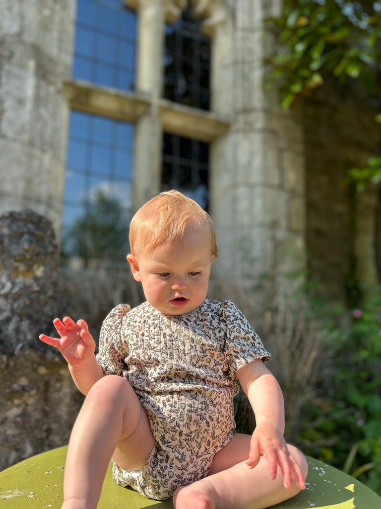 baby sat in sunny garden in front of old stone house wearing short sleeved cream ribbed jersey onesie with bird and flower print