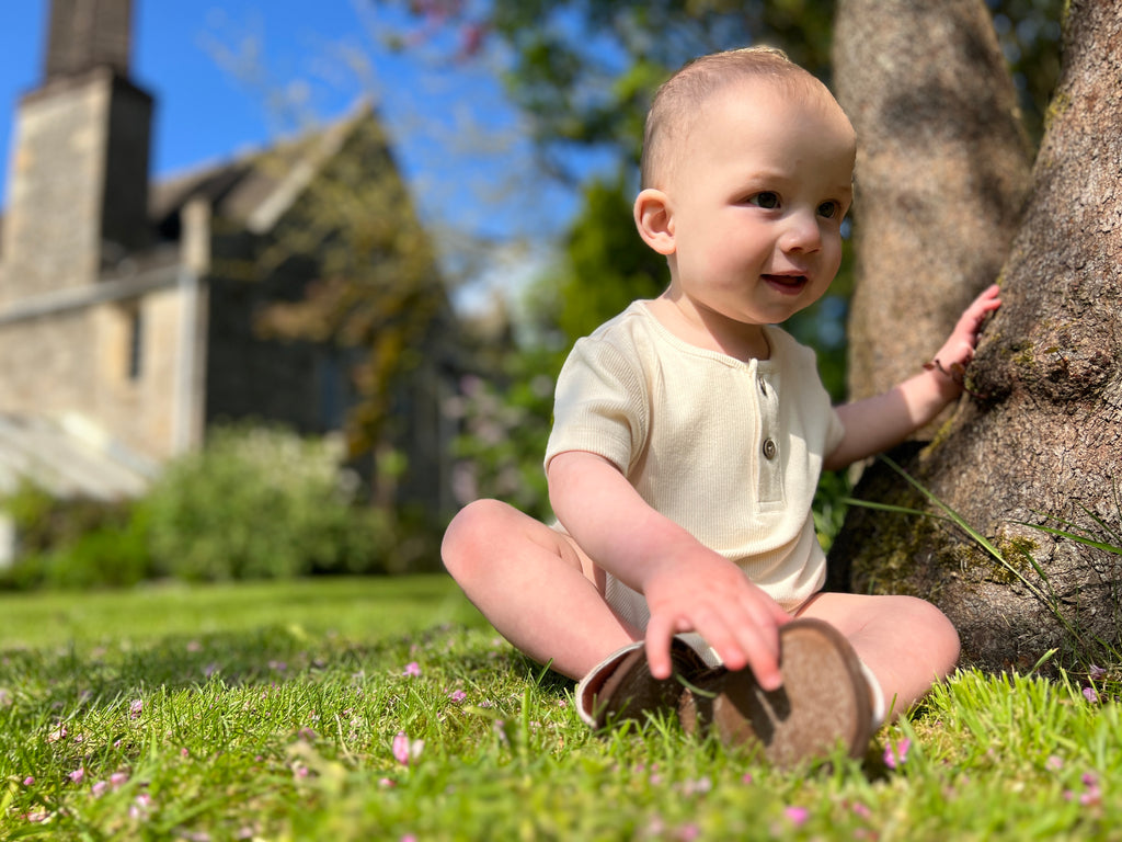 happy baby by a tree on sunny day wearing short sleeved cream onesie with 2 button henley style detail on front