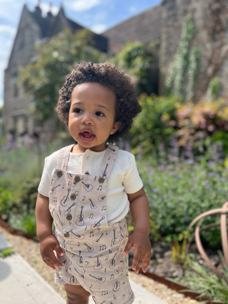 toddler stood in front of old stone house in sunny garden wearing cream overalls with buttons down the front 2 patch pockets and guitar print all over