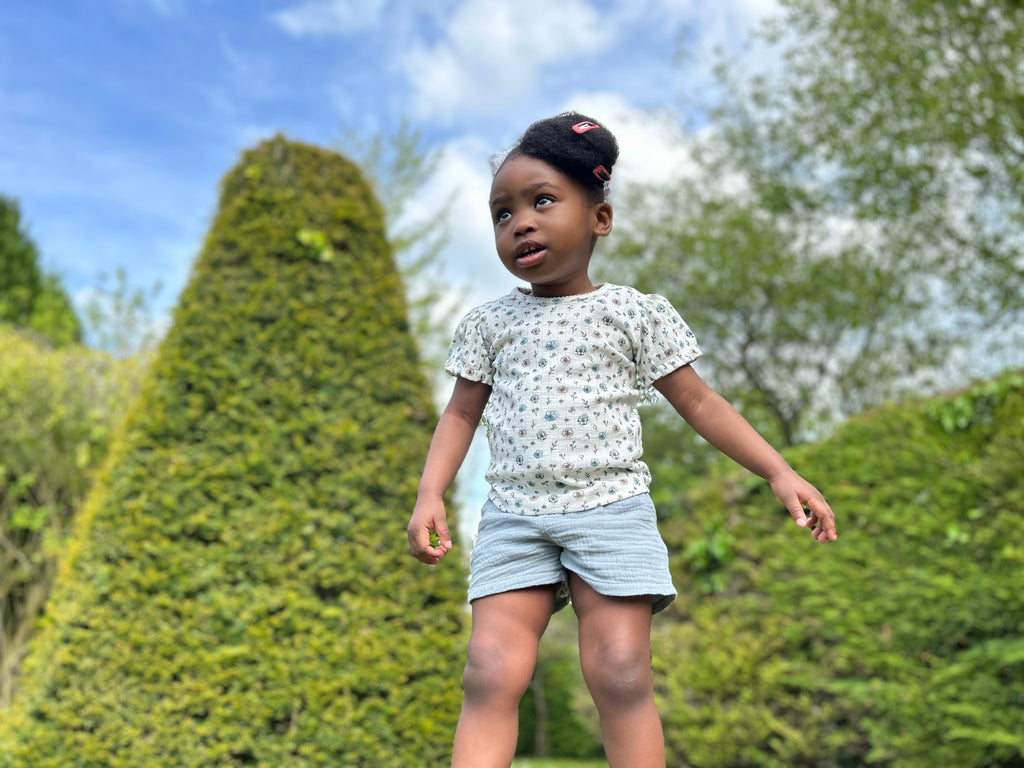 girl in garden standing in front of green hedges wearing tee shirt in white ribbed jersey with flower print and blue shorts
