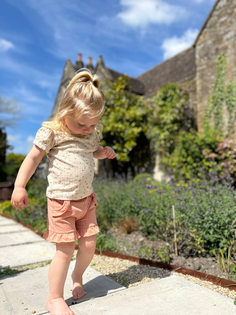 girl walking on path in sunny garden wearing coral shorts in jersey pointelle fabric side pockets and frill around the legs and cream tee