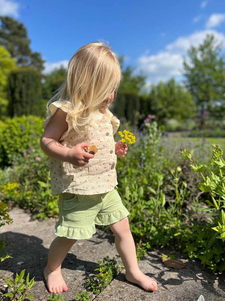 happy toddler walking on old stone path holding summer flowers on sunny day wearing frill sleeved blouse with square cut neck line and buttons down the front and green gauze shorts