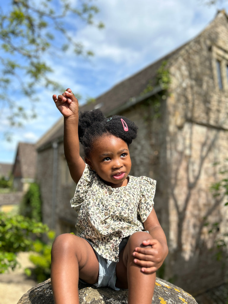 toddler sat on old stone on sunny day wearing frill sleeved blouse with flower and bird small print square cut neck line and buttons down the front