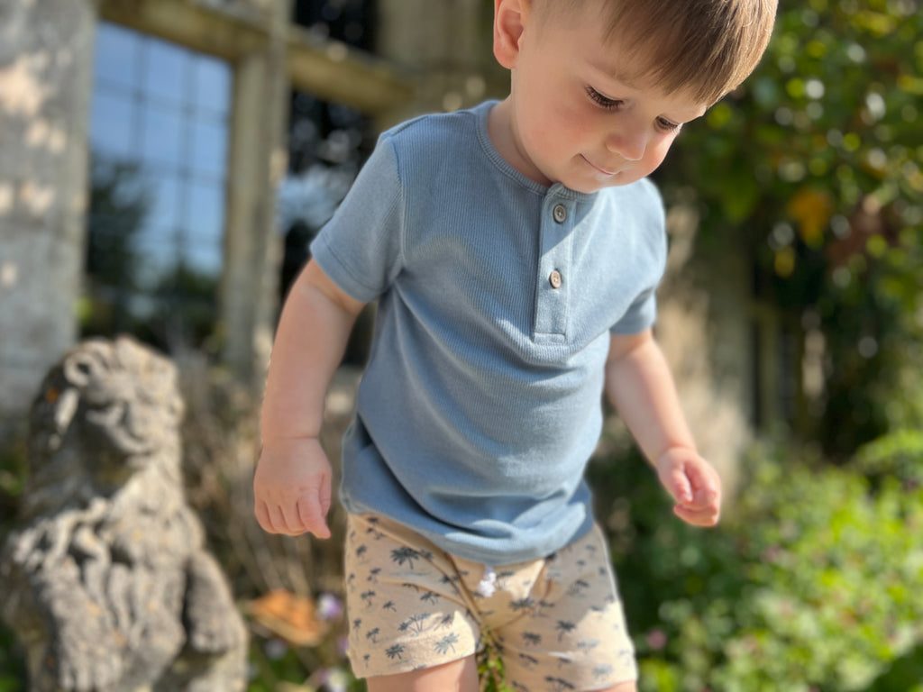 happy toddler in sunny garden wearing pale blue short sleeved henley tee with 2 button detail and island print shorts