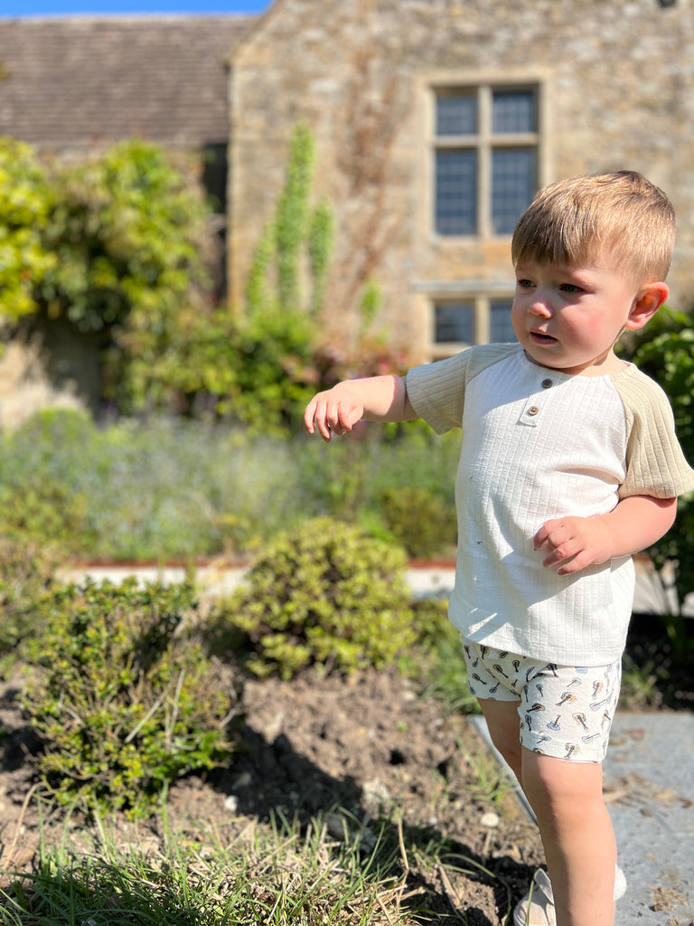 toddler standing in front of old stone manor on sunny day wearing white jersey shorts with drawstring waist all over guitar tree print and white tee shirt