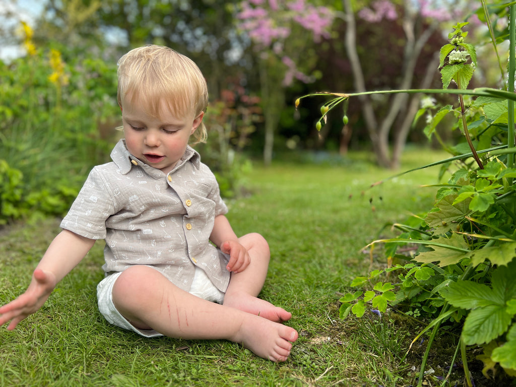 toddler sat in sunny garden wearing short sleeved grey gauze shirt front chest pocket buttons all the way down front  and white shorts