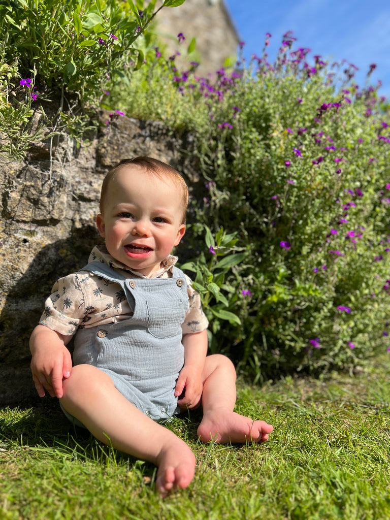 happy baby sat in sunny garden in front of summer flowers wearing blue shortie gauze overalls