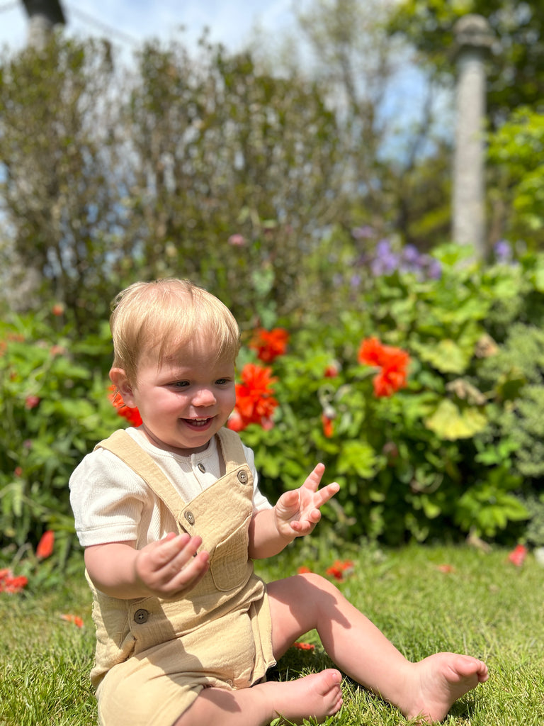 happy toddler sat in sunny garden in front of summer flowers wearing gold shortie gauze overalls 