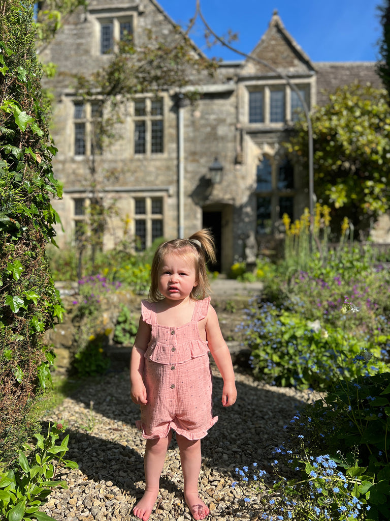 girl standing in front of old stone house in garden on sunny day wearing pink shortie overalls with frill shoulder straps and buttons down the front 