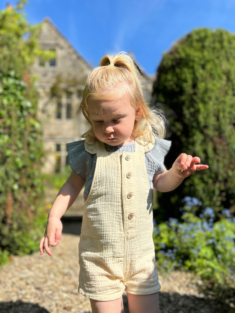 toddler in sunny garden wearing blue gauze blouse with capped sleeves buttons down the front 2 patch pockets on front seahorse print all over under cream overalls