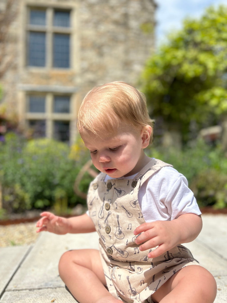 toddler sat in front of old stone house in sunny garden wearing cream overalls with buttons down the front 2 patch pockets and guitar print all over