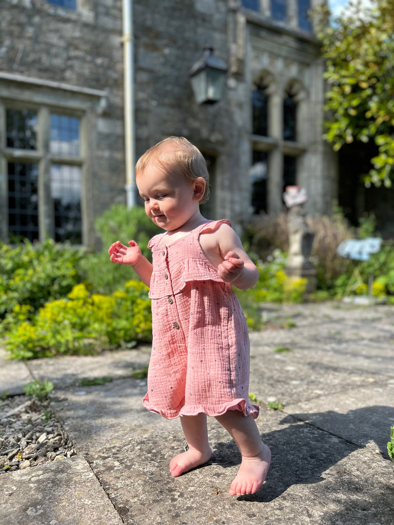 happy toddler standing in front of old stone manor house wearing  pale pink gauze shortie overal all over dotty print. buttons on front and frill detail round front and legs