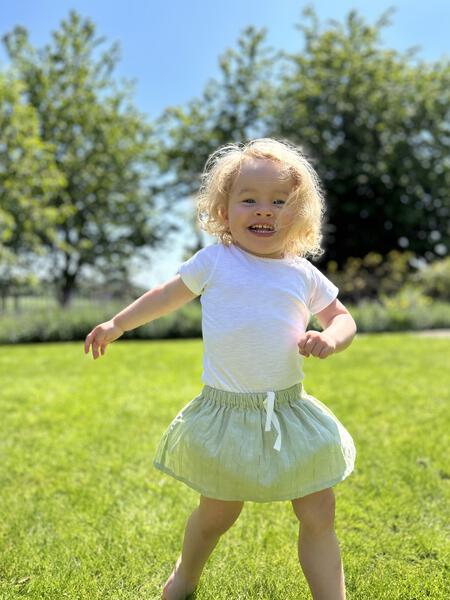 happy toddler in sunny garden wearing white onesie with poppers at side on neck opening and green skirt