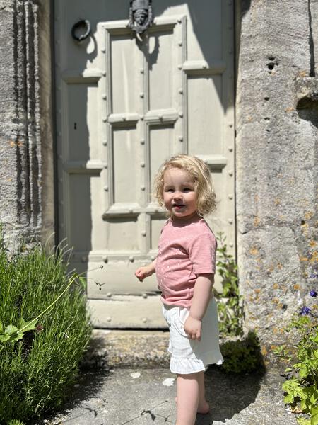 happy toddler standing in front of old wooden front door wearing pink short sleeve tee with heart dot print and white shorts