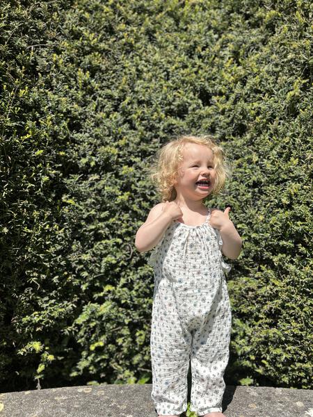 happy toddler standing infront of green garden hedage in summer sunshine wearing jersey long legged romper with shoe string straps and frills Small flowers print on white background