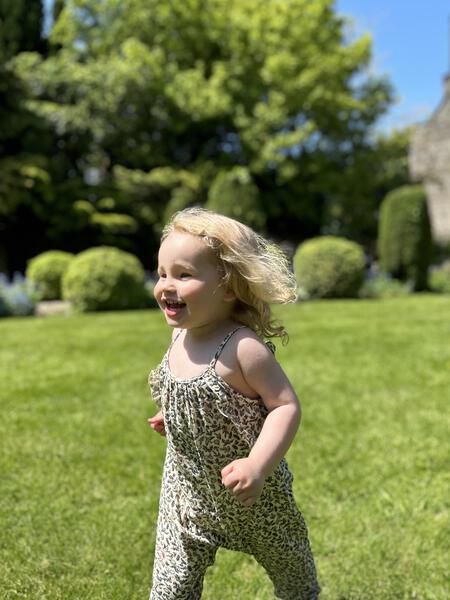 happy toddler running in sunny garden wearing  jersey long legged romper with shoe string straps and frills Small flowers and birds print on cream background