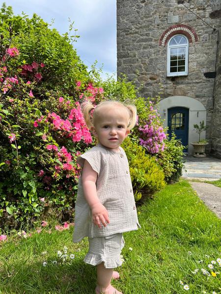 toddler in sunny garden standing in front of stone house wearing grey gauze shorts with 2 side pockets and frill around legs and grey blouse