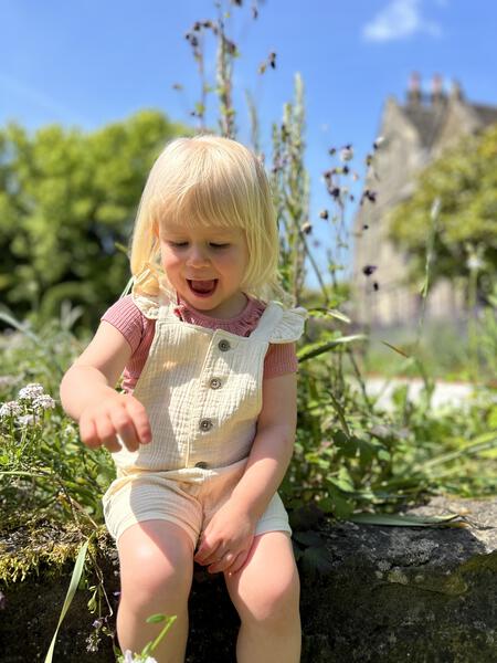 toddler in sunny garden sat on stone wall and wearing pink tee shirt with patterned fabric and frill neck under cream overalls