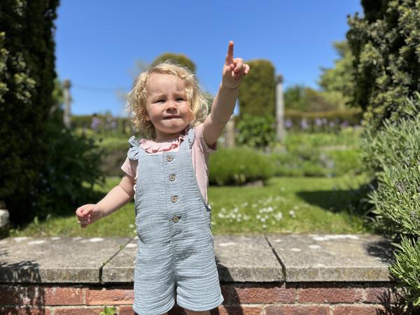 toddler in sunny garden pointing and wearing pink tee shirt with patterned fabric and frill neck under blue overalls