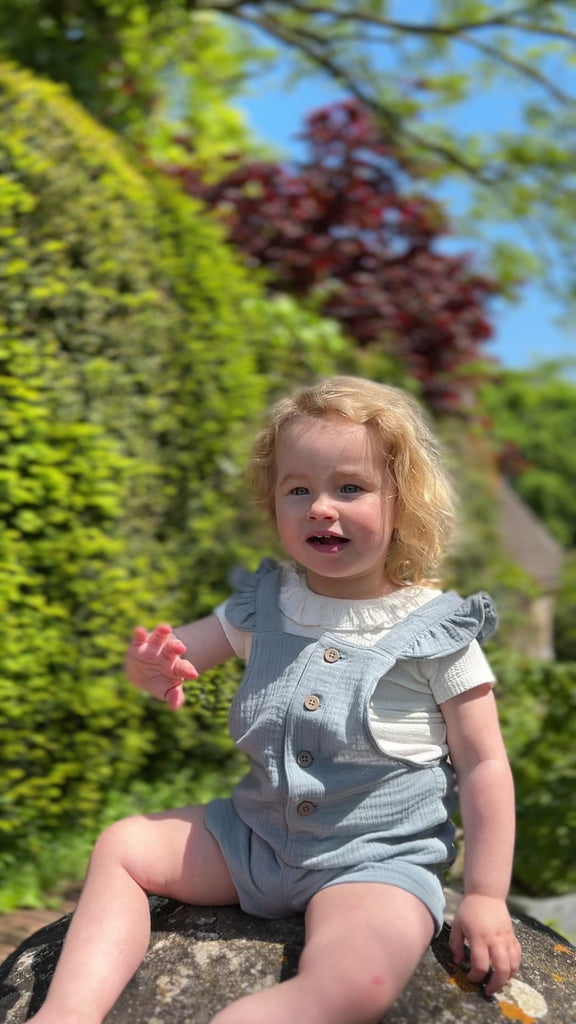 happy toddler sat in sunny garden waving wearing cream tee shirt with patterned fabric and frill neck under blue overalls