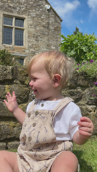 happy toddler sat in front of old stone house in sunny garden wearing cream overalls with buttons down the front 2 patch pockets and guitar print all over