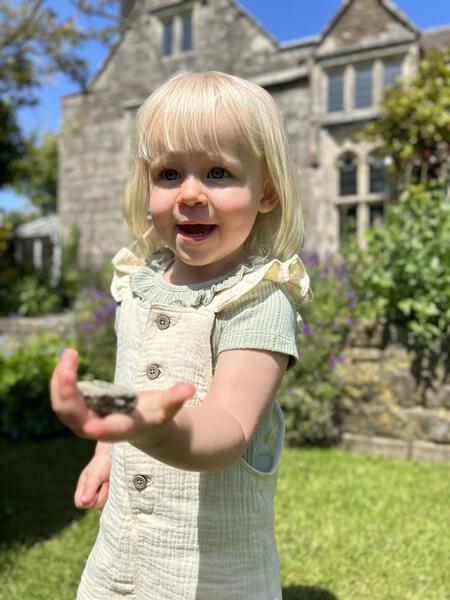toddler in sunny garden wearing green tee shirt with patterned fabric and frill neck under cream overalls