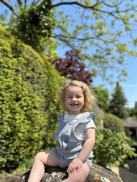 toddler in sunny garden sat on stone wall and wearing white tee shirt with patterned fabric and frill neck under blue overalls