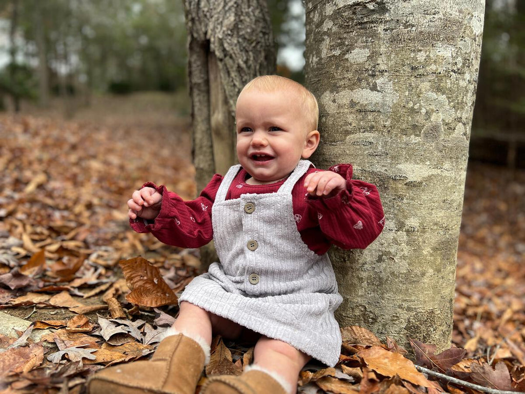 baby sat in autumn wood next to treee wearing burgundy long sleeve onesies small print winter scene items ice skates, mittens, pompom hat pinafore dress in jumbo cord