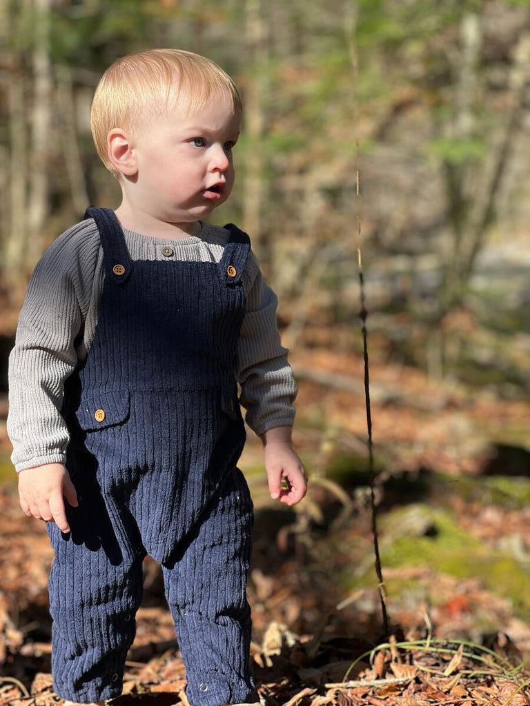 toddler standing in autumn woodland wearing dark grey onesie with 3 button detail on the front slight gathered neck line under navy cord overalls