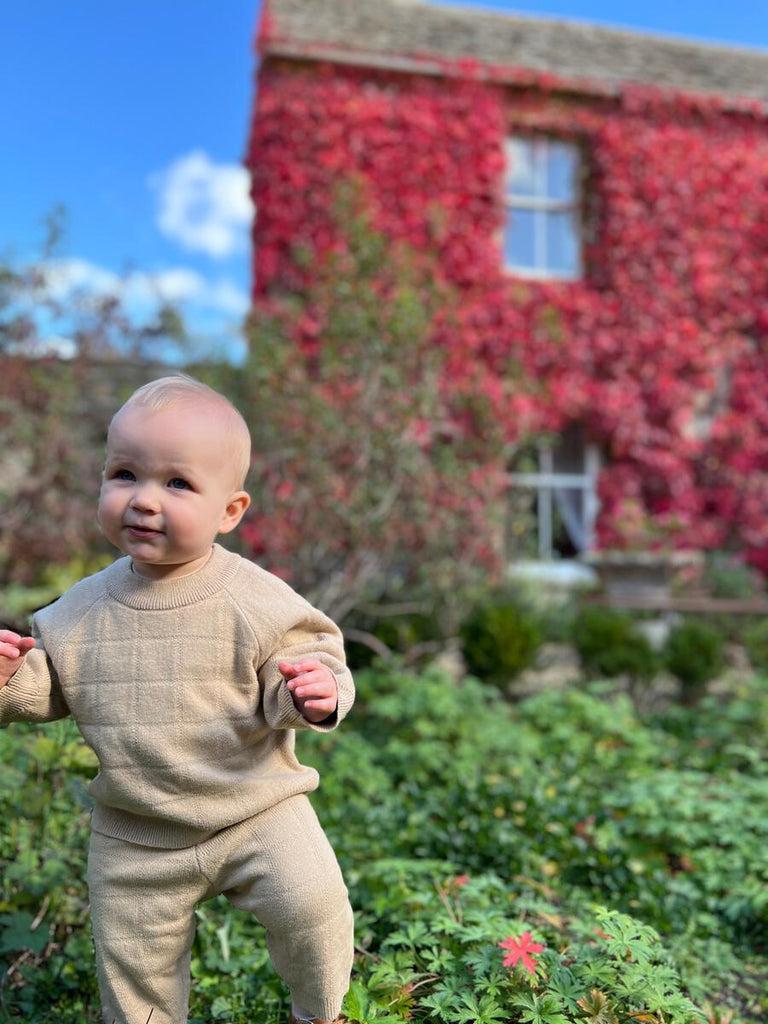 happy baby stood in front of red autumn leaves wearing oatmeal knit sweater with square pattern and matching pants