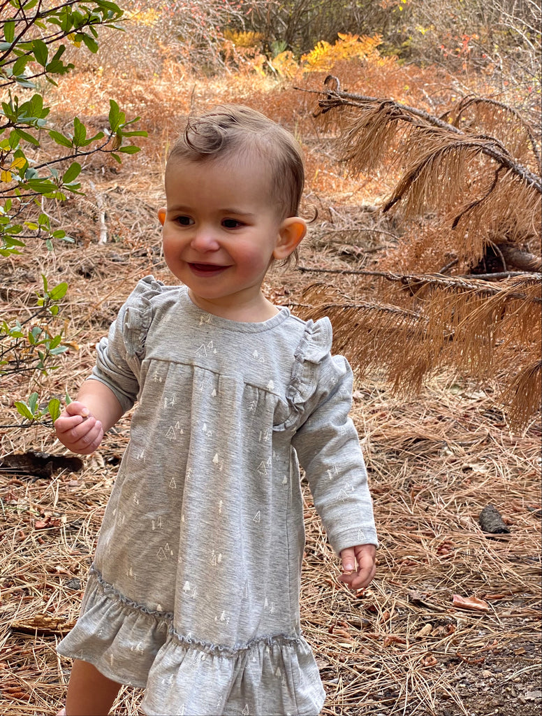 happy toddler in autumn wood wearing pale grey dress with tree print with ruffle shoulders and long sleeves ruffle frill around the bottom