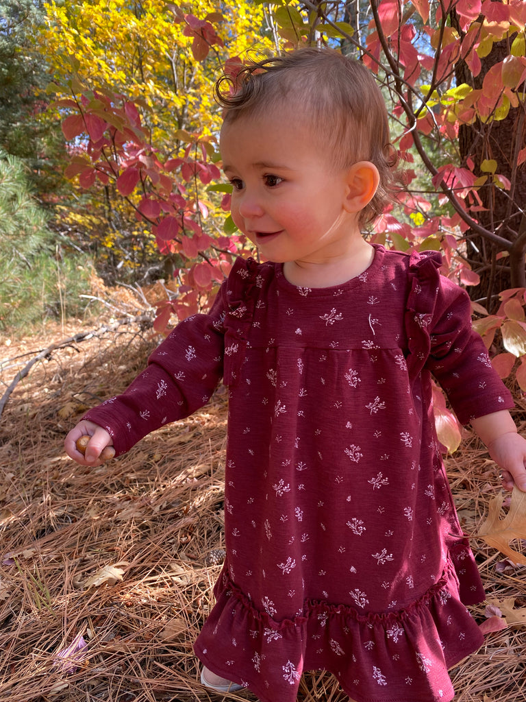 toddler standing smiling in autumn wood wearing burgundy dress with ruffle shoulders and long sleeves ruffle frill around the bottom small flower print in white all over