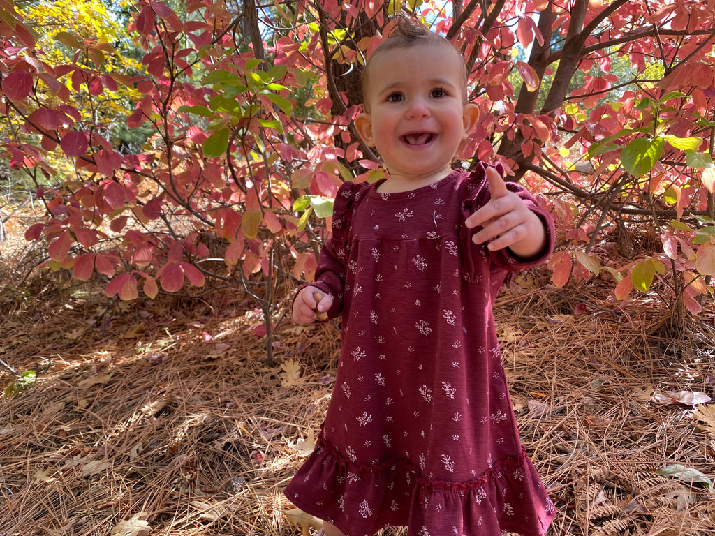toddler standing smiling in autumn wood wearing burgundy dress with ruffle shoulders and long sleeves ruffle frill around the bottom small flower print in white all over