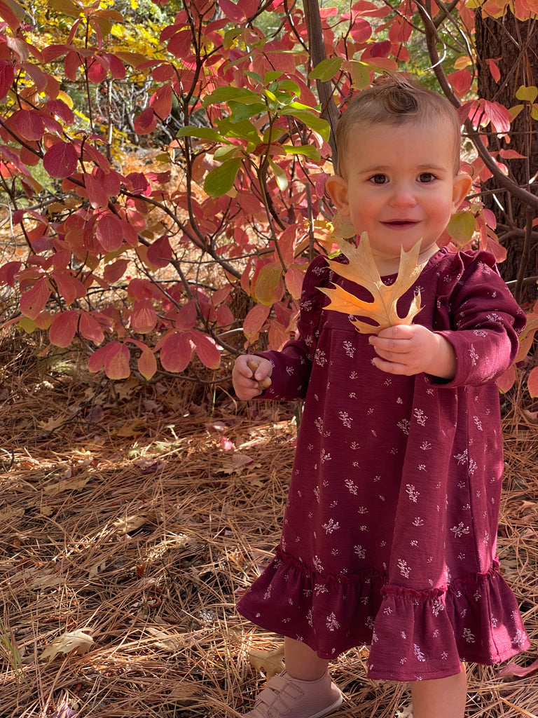 toddler standing smiling holding leaf in autumn wood wearing burgundy dress with ruffle shoulders and long sleeves ruffle frill around the bottom small flower print in white all over