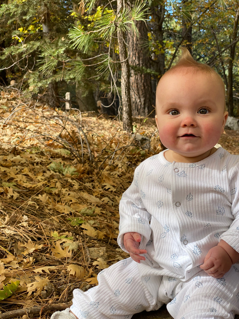 happy baby sat in autumn wood wearing white ribbed romper with leaf print. poppers down front