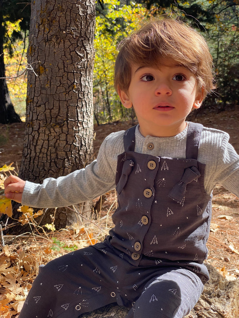 happy toddler sat in sunny autumn woods wearing grey overalls with rocket all over print. knotted straps and buttons down the front bib. patch pockets at side and grey ribbed tee underneath