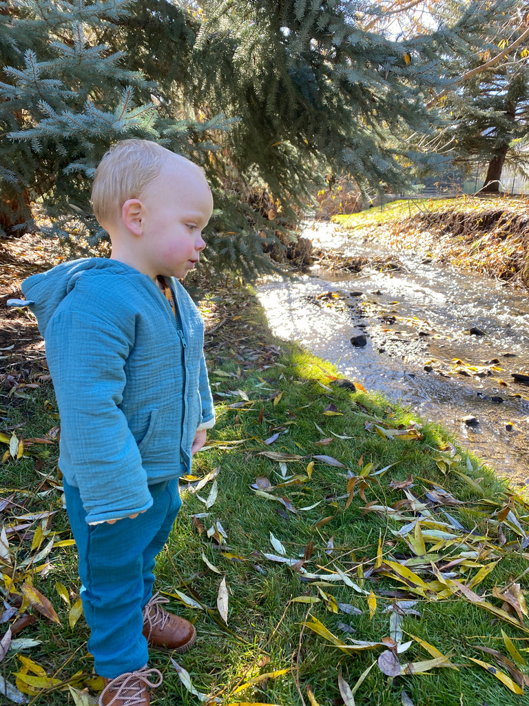 toddler standing in autumnal wood near stream wearing padded jacket zip front with hood teal green jersey with leaf print lining