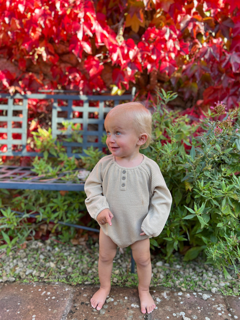 toddler standing in fromnt of red autumn leaves wearing oatmeal onesie with 3 button detail on the front slight gathered neck line 