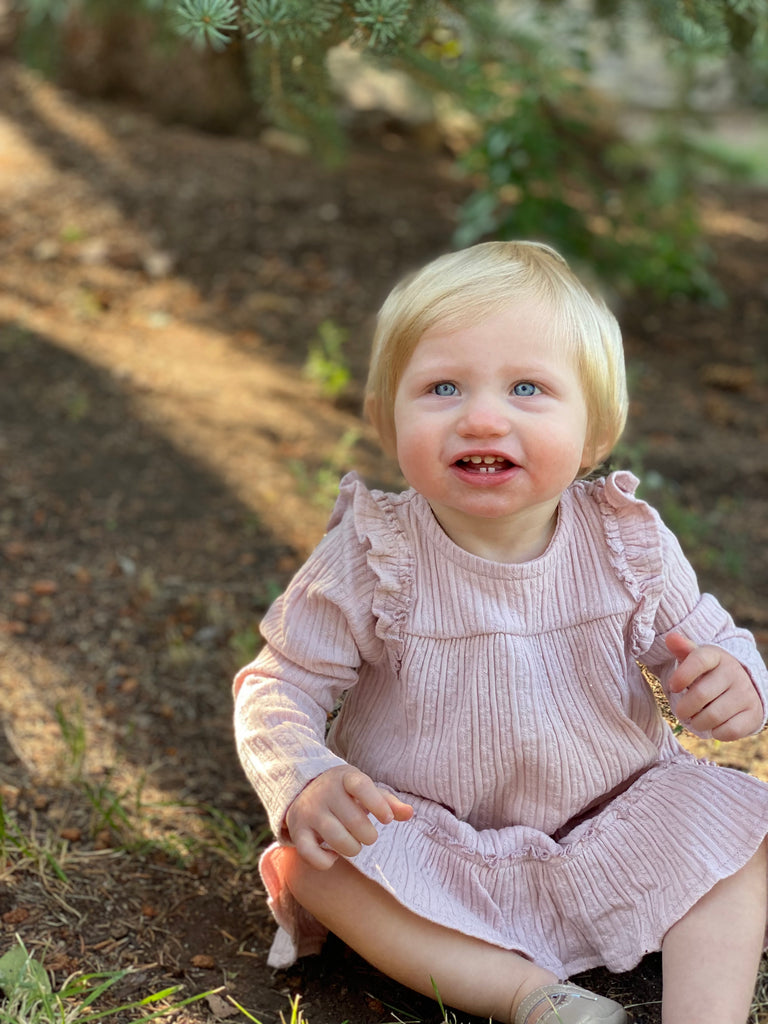 happy toddler sitting in autumn wood wearing pink dress with ruffle shoulders and long sleeves ruffle frill around the bottom