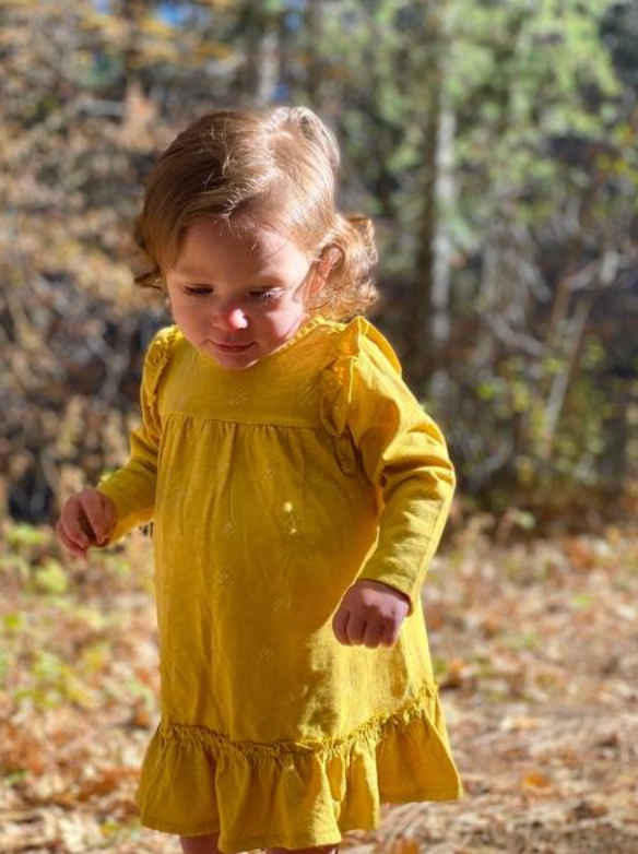 toddler in sunny autumn wood wearing bright yellow dress with ruffle shoulders and long sleeves ruffle frill around the bottom  arrow print all over