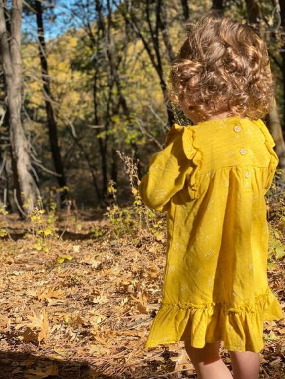 back view of toddler in sunny autumn wood wearing bright yellow dress with ruffle shoulders and long sleeves ruffle frill around the bottom arrow print all over
