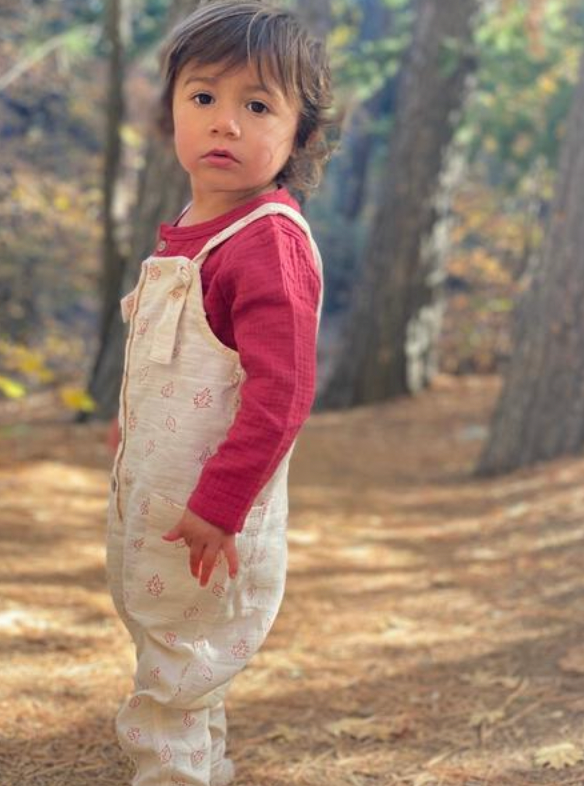 toddler standing in sunny autumn woods wearing cream overalls with leaf all over print. knotted straps and buttons down the front bib. patch pockets at side and red gauze top underneath