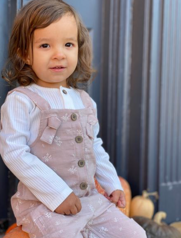 happy toddler sat on pumpkins wearing pink overalls with flower all over print. knotted straps and buttons down the front bib. patch pockets at side white rib tee underneath